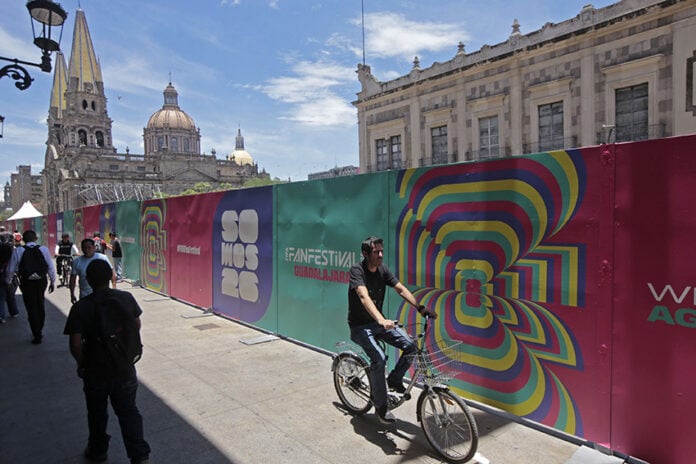 Pedestrians pass a colorful fence with World Cup messaging, with the Guadalajara cathedral in the background