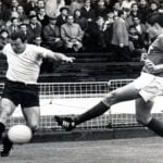 Two national selection team members for Uruguay and Mexico battle on the soccer pitch for control of the ball during a game at Wembley Stadium. Fans, mainly men and boys, in trenchcoats, watch the game in the bleachers.