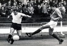 Two national selection team members for Uruguay and Mexico battle on the soccer pitch for control of the ball during a game at Wembley Stadium. Fans, mainly men and boys, in trenchcoats, watch the game in the bleachers.