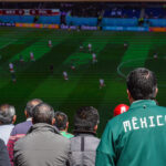 A man wearing a Mexico soccer jacket watches a World Cup match on a screen in a crowd