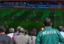A man wearing a Mexico soccer jacket watches a World Cup match on a screen in a crowd