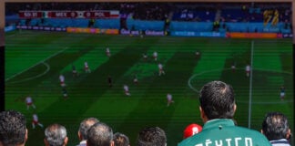 A man wearing a Mexico soccer jacket watches a World Cup match on a screen in a crowd