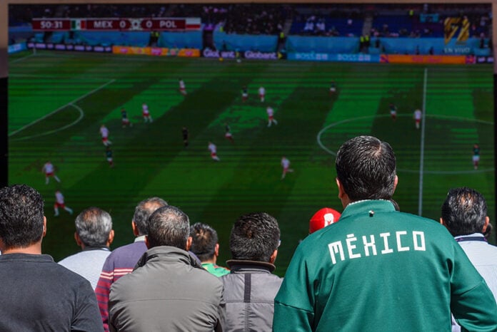 A man wearing a Mexico soccer jacket watches a World Cup match on a screen in a crowd
