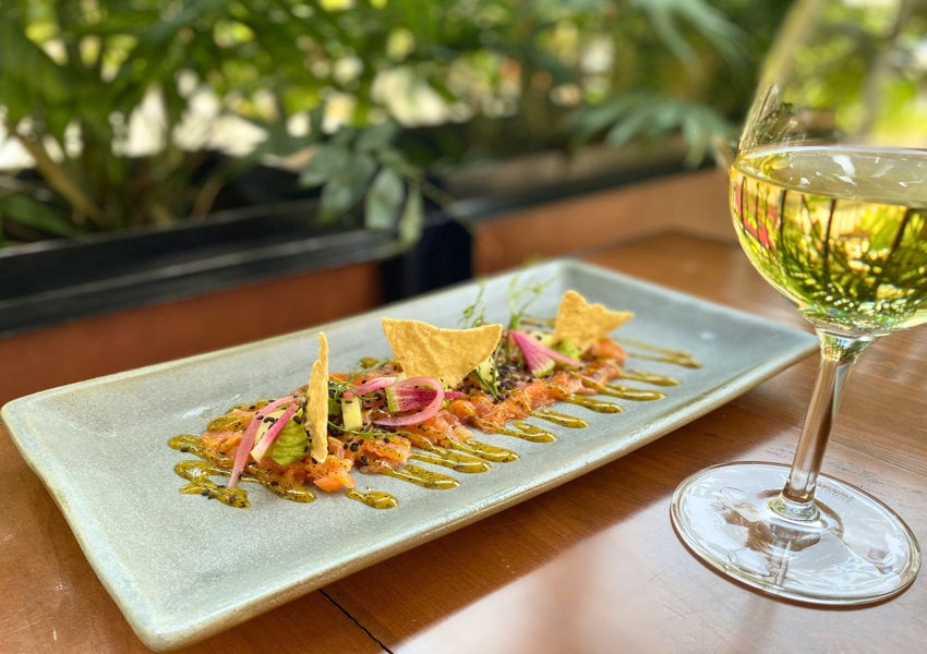 A haute cuisine presentation of ceviche toppped with red onions and tortilla chips on a gray ceramic rectangular plate, on a wooden table. A glass of white white is next to the plate.