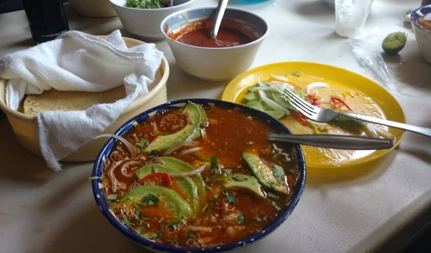A bowl of Mexican birria on a white plastic table. Beside it are a half eaten plate of vegetables, a bowl with a white napkin and tortillas inside.