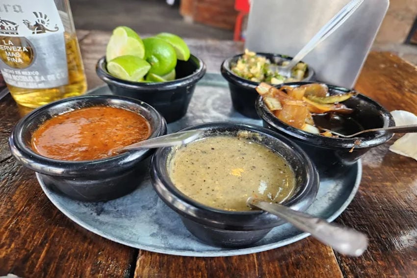 A simple melamine plate holding small bowls filled with salsa, lime slices, guacamole, and grilled onions, the typical fixings available at taquerias in Mexico City and near Estadio Azteca.