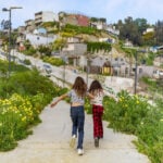 Two Mexican girls seen from behind running through pathway in Xicoténcatl Park on the outskirts of Tijuana, Mexico. In the background is a community built on a hillside of cheaply made, in some cases unfinished concrete homes.