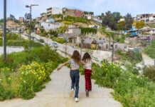 Two Mexican girls seen from behind running through pathway in Xicoténcatl Park on the outskirts of Tijuana, Mexico. In the background is a community built on a hillside of cheaply made, in some cases unfinished concrete homes.