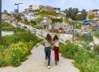 Two Mexican girls seen from behind running through pathway in Xicoténcatl Park on the outskirts of Tijuana, Mexico. In the background is a community built on a hillside of cheaply made, in some cases unfinished concrete homes.