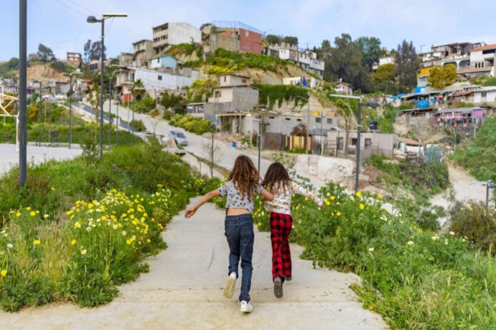 Two Mexican girls seen from behind running through pathway in Xicoténcatl Park on the outskirts of Tijuana, Mexico. In the background is a community built on a hillside of cheaply made, in some cases unfinished concrete homes.