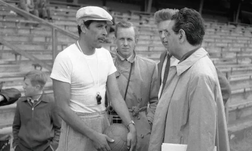 Legendary Mexican soccer coach Ignacio Trelles in a white men's tee and sweatpants, holding a soccer ball by his hips as he converses with three unidentified men in trenchcoats near the nearly empty bleachers of a sports stadium.