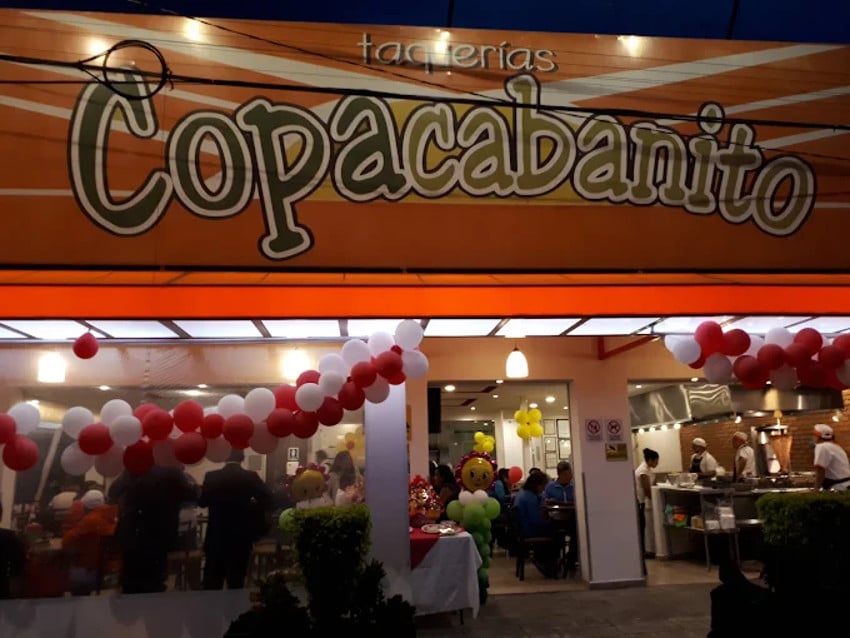 A taqueria in Mexico City near Estadio Azteca with a large sign saying "Taquerias Copacabanito." It features outdoor seating and an open kitchen with chefs