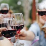 Multiple hands of people seated at a table. They are holding wine glasses with red wine and reaching out to toast. The photo is against a bokeh background