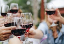 Multiple hands of people seated at a table. They are holding wine glasses with red wine and reaching out to toast. The photo is against a bokeh background