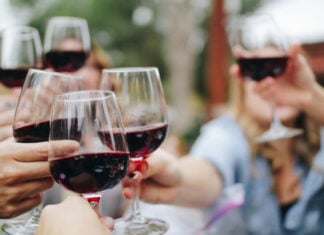 Multiple hands of people seated at a table. They are holding wine glasses with red wine and reaching out to toast. The photo is against a bokeh background