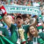 A gray-haired man waves a "Mexico" banner in a soccer stadium surrounded by other fans