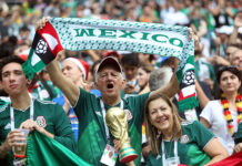 A gray-haired man waves a "Mexico" banner in a soccer stadium surrounded by other fans