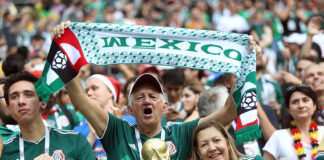 A gray-haired man waves a "Mexico" banner in a soccer stadium surrounded by other fans