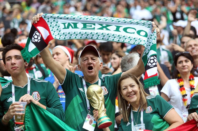 A gray-haired man waves a "Mexico" banner in a soccer stadium surrounded by other fans