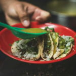 Tacos on a red plate with a hand holding a plastic tablespoon and using it to pour green salsa on the three tacos.