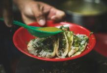 Tacos on a red plate with a hand holding a plastic tablespoon and using it to pour green salsa on the three tacos.