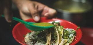 Tacos on a red plate with a hand holding a plastic tablespoon and using it to pour green salsa on the three tacos.