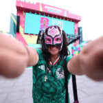 A young woman standing outside in a Guadalajara plaza wearing a Mexico National Selection T-shirt and a lucha libre mask over most of her face poses, smiling, with arms outstretched toward the camera. In the distant background, the colorful FIFA Fan Fest event stage can be seen.