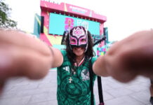A young woman standing outside in a Guadalajara plaza wearing a Mexico National Selection T-shirt and a lucha libre mask over most of her face poses, smiling, with arms outstretched toward the camera. In the distant background, the colorful FIFA Fan Fest event stage can be seen.