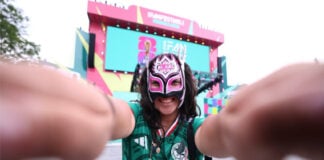 A young woman standing outside in a Guadalajara plaza wearing a Mexico National Selection T-shirt and a lucha libre mask over most of her face poses, smiling, with arms outstretched toward the camera. In the distant background, the colorful FIFA Fan Fest event stage can be seen.