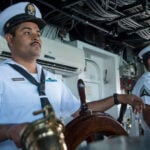 Two uniformed Mexican navy personnel at the helm of a Mexican navy ship. One man has his hand resting on the ship's steering wheel