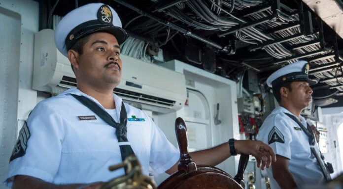 Two uniformed Mexican navy personnel at the helm of a Mexican navy ship. One man has his hand resting on the ship's steering wheel