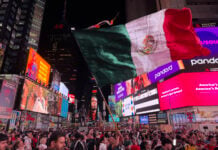 A crowd of fans filling 42nd Street in Times Square, New York City, surrounded by the buildings and giant digital screens Times Square is known for. One fan in the center of the photo is waving a huge Mexican flag.