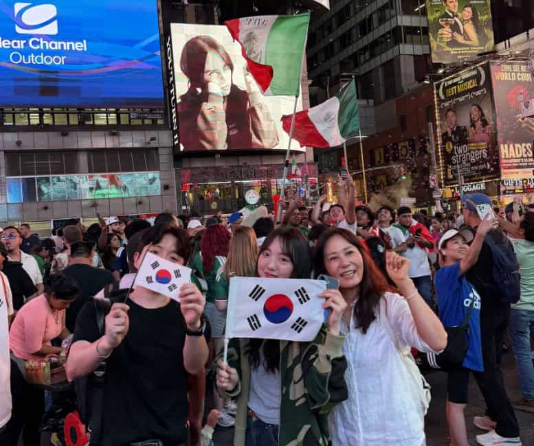 Korea World Cup soccer fans in Times Square New York. Three Korea fans holding small South Korean flags pose for the camera. In the background are skyscrapers and advertising billboards typical to Times Square, as well as a Mexico team fans holding different large Mexican flags.