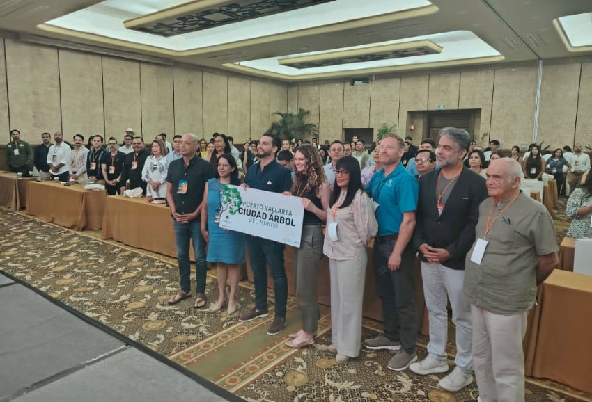 Right Puerto Vallarta people in casual clothing posing for a formal photo in a conference room while many people behind them look on. The eight people are holding up a small banner that says in Spanish: Puerto Vallarta Ciudad Arbol del Mundo.