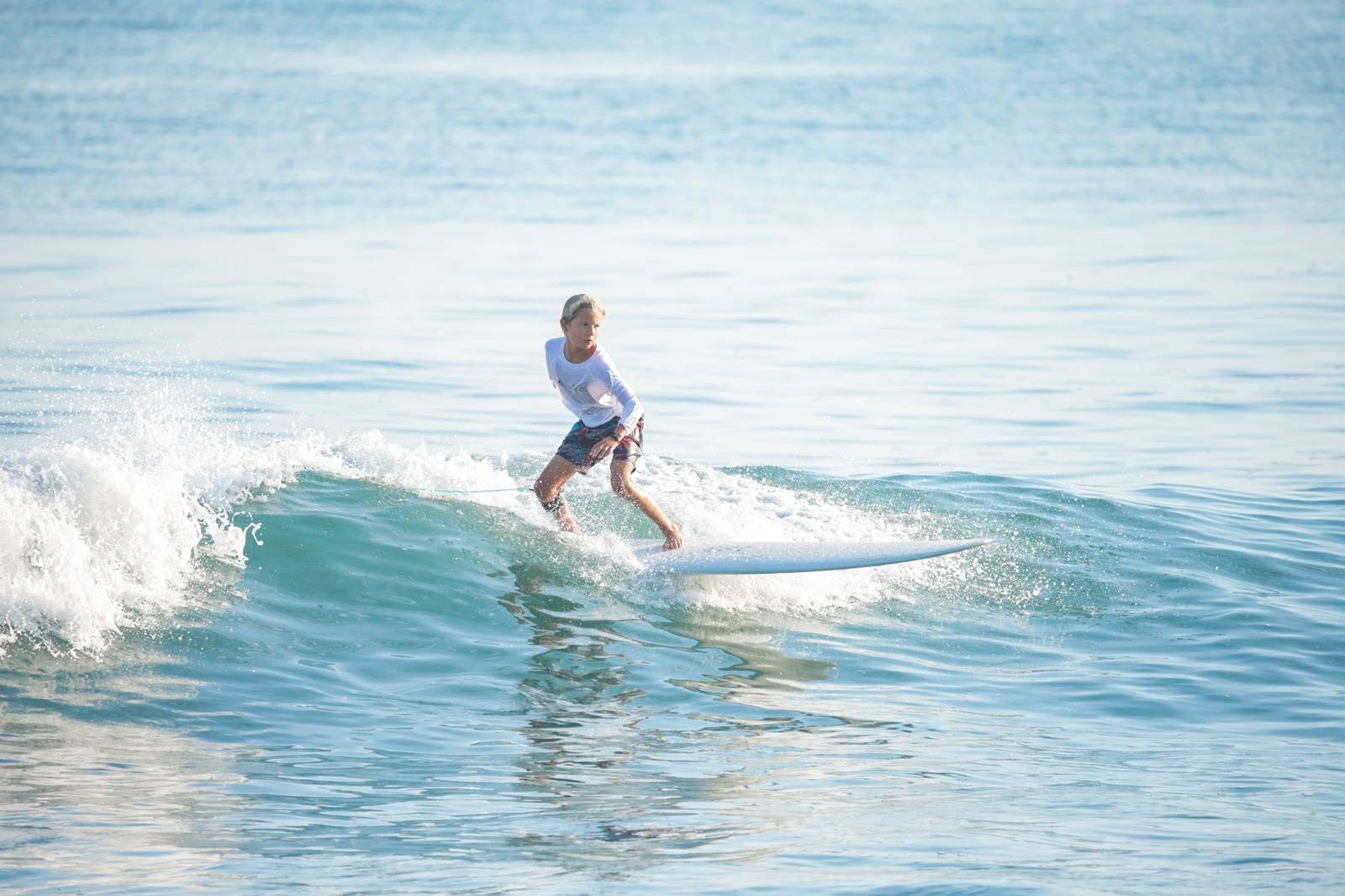 A young woman with short blonde hair dressed in a white longsleeved shirt and bermuda shorts surfs an ocean tidal wave in Mexico toward the shore.