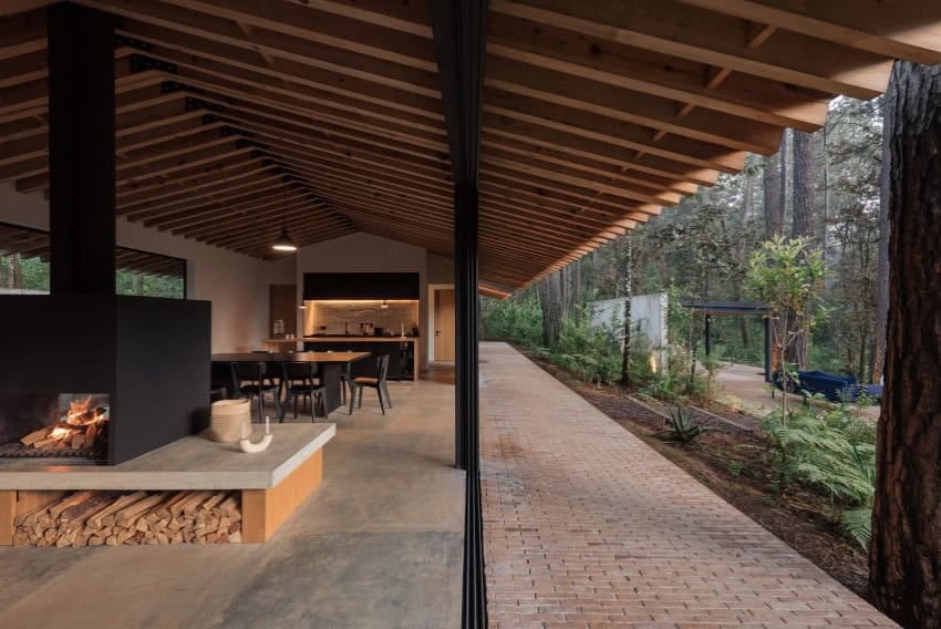 A wide, brick tiled with a gabled roof sits in a forest environment surrounded by tall pines. The patio has a wood stove, a handmade luxury wooden table and an outdoor bar.