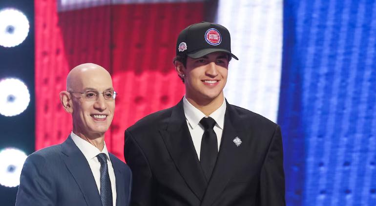 A young clean cut man in a black suit and tie and white oxford shirt and wearng a baseball cap with a red circular path on the front smiles and poses for a picture with an older bald man wearing glasses, also in a suit and tie in dark blue and a white oxford shirt. They are posing at some at an NBA press conference.