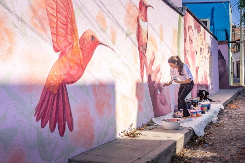 A female artist in the historic Mexican town of San Miguel de Allende paints a vibrant pink and orange hummingbird mural on a concrete street wall as she stands on on a white tarp laid out on the sidewalk where several paint cans are arranged.
