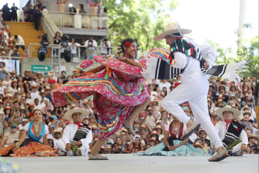 A pair of dancers in a traditional, colorful dress and white suit twirl on a stage in front of hundreds of people at Guelaguetza in Oaxaca.