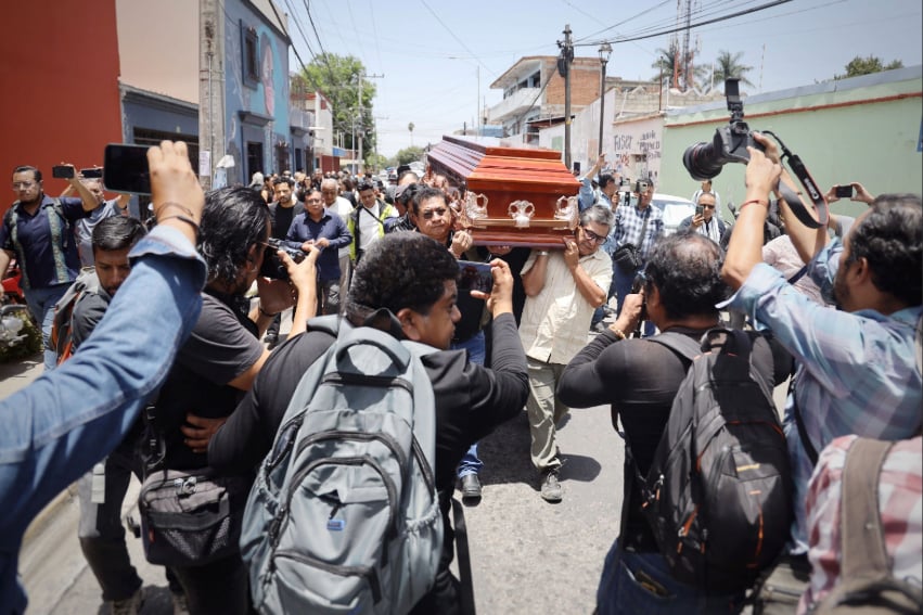 Dozens of photographers take pictures as a casket is carried down a street in Oaxaca city