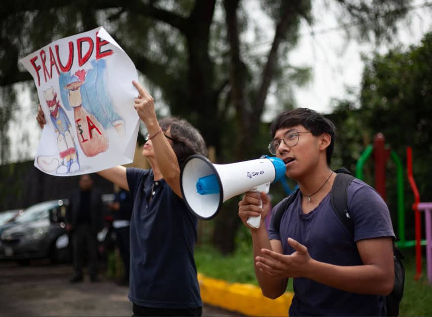 Students yell into a megaphone, with one carrying a sign reading "Fraude"