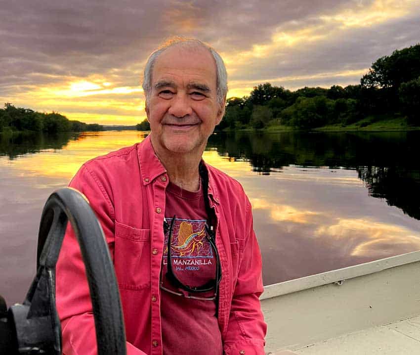 an elderly man smiling at camera while on a river wearing a red shirt and t-shirt.