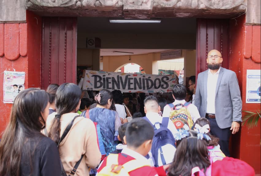 A teacher stands with a welcome banner as students flood into a Cuernavaca public school