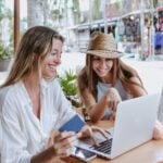 young latin women friends using laptop to shopping with credit card at restaurant at beach in Mexico Latin America, hispanic female in caribbean city with tropical weather.