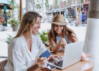 young latin women friends using laptop to shopping with credit card at restaurant at beach in Mexico Latin America, hispanic female in caribbean city with tropical weather.