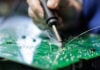 Closeup of technician soldering on a green PCB with visible flux smoke during electronics assembly at a factory workstation