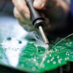 Closeup of technician soldering on a green PCB with visible flux smoke during electronics assembly at a factory workstation