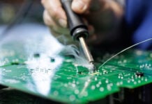 Closeup of technician soldering on a green PCB with visible flux smoke during electronics assembly at a factory workstation