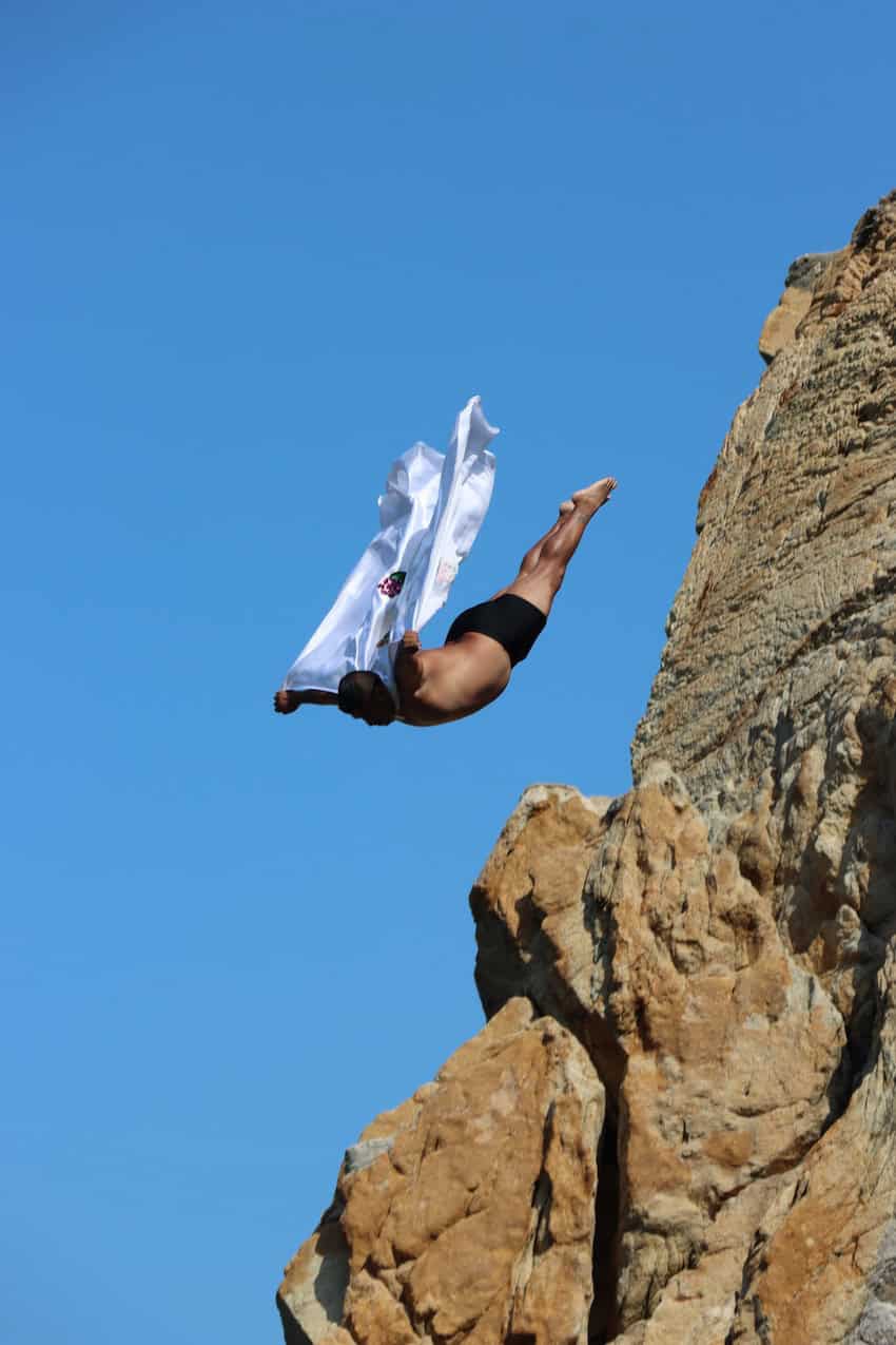 A cliff diver wearing a white cape dives forwards into the ocean