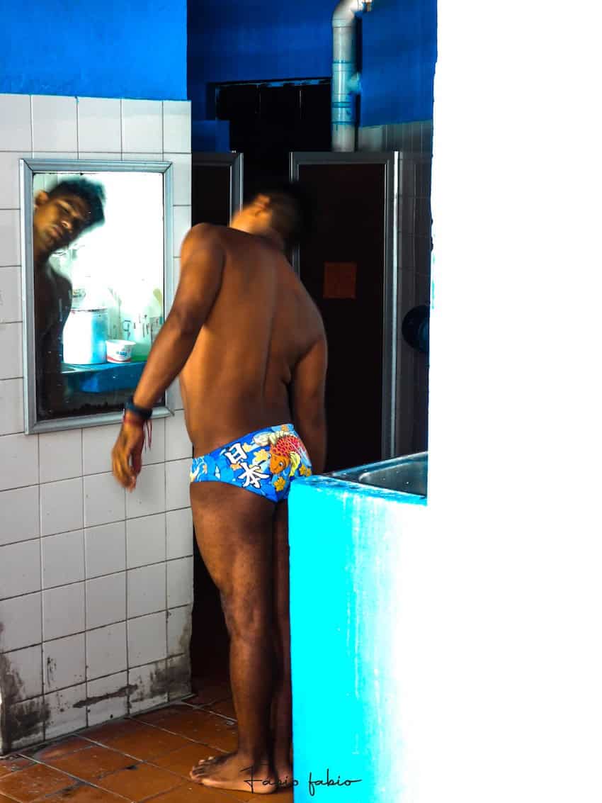 A male cliff diver stretches his neck in front of a mirror in a changing room.